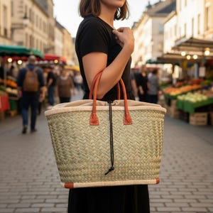 May include: Woven straw tote bag with tan leather handles and accents. The bag has a beige fabric lining and a drawstring closure. The bag is rectangular and appears to be sturdy, suitable for carrying groceries or other items. The bag is being held by a person in a black dress.