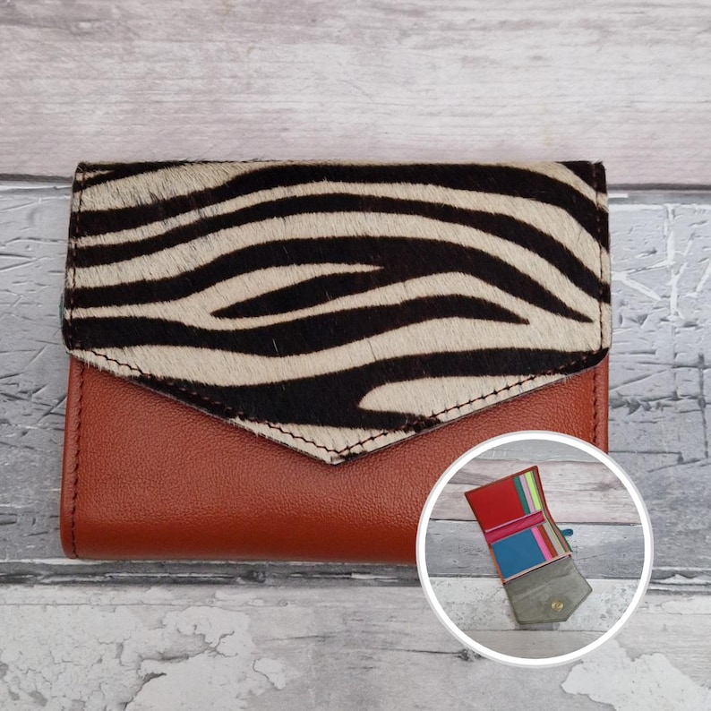 May include: A brown leather wallet with a zebra print flap. The wallet has a bi-fold design and a small, circular inset showing the open interior with colourful compartments. The wallet is closed in the image.