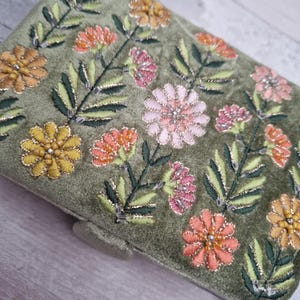 May include: Olive green velvet clutch with embroidered floral design. The flowers are in shades of yellow, orange, and pink, with green leaves and gold accents. The clutch has a rectangular shape.