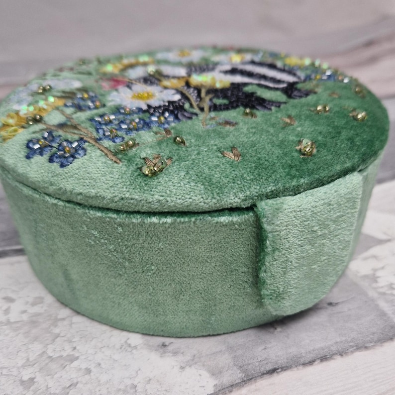 May include: A round, light green velvet jewelry box with a detailed embroidered lid. The lid features a scene with white daisies, blue flowers, and a badger. The box has a matching green side and a soft, plush texture.