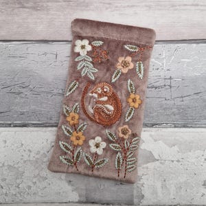 Puede incluir: Funda de gafas de terciopelo malva con un diseño bordado. El diseño presenta una ardilla enroscada rodeada de flores en tonos naranja, blanco y verde. La funda es rectangular con la parte superior redondeada.