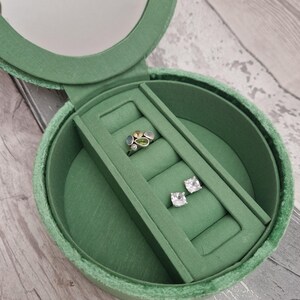May include: Open green velvet jewelry box with a mirror in the lid. Inside, a silver ring with colorful stones and a pair of square-cut, clear stone earrings are displayed on a green velvet insert. The box is round.