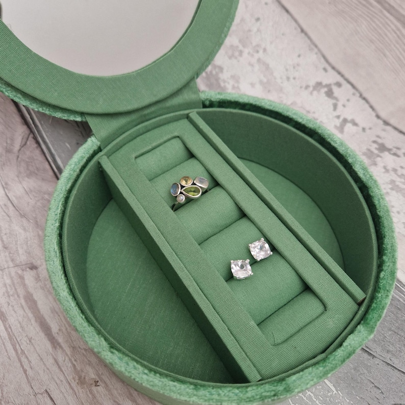 May include: Open green velvet jewelry box with a mirror in the lid. Inside, a silver ring with colorful stones and a pair of square-cut, clear stone earrings are displayed on a green velvet insert. The box is round.