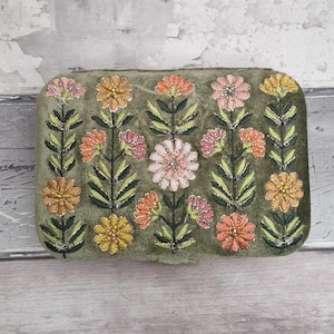 May include: A rectangular, olive green velvet clutch with an embroidered floral design. The flowers are in shades of pink, orange, and yellow, with green leaves and stems. The embroidery is detailed and adds texture to the clutch.