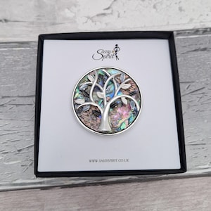 May include: A circular brooch featuring a silver-toned tree silhouette layered over an iridescent abalone shell inlay. The piece is presented in a white square box with the text Sassy Spirit and the website URL WWW.SASSYSPIRIT.CO.UK printed on it.