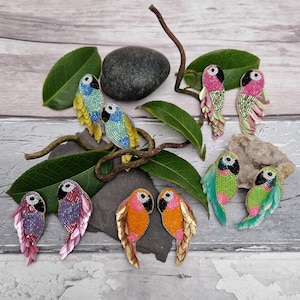 May include: A selection of beaded parrot brooches in shades of blue, pink, orange, and green. The brooches are displayed on a wooden surface with leaves and a stone, highlighting their detailed craftsmanship and bright colours.