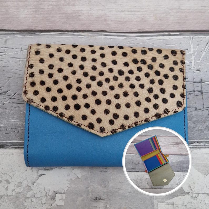 May include: A blue leather wallet with a flap closure. The flap is made of tan-coloured animal hide with black polka dots. A smaller, open wallet with colourful stripes is visible in the lower right corner.