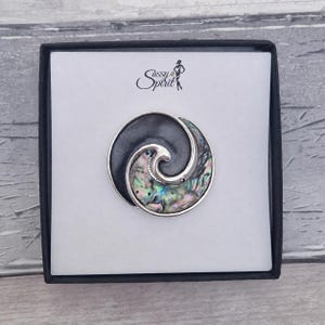 May include: A circular brooch featuring a spiral design with a polished dark grey section and an iridescent abalone shell inlay. The piece is set in a silver-toned metal frame and presented on a white display card with the text Sassy Spirit.