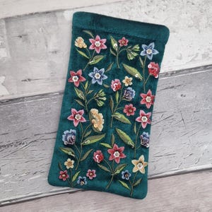 Könnte beinhalten: Ein teal-farbenes Brillenetui aus Samt mit einem gestickten Blumendesign. Die Blumen sind in Rosa-, Blau-, Gelb- und Weißtönen gehalten, mit grünen Blättern und Stielen. Das Etui ist rechteckig mit abgerundeter Oberseite.