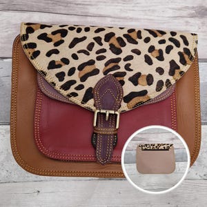 May include: A multi-colored leather handbag featuring a tan base, a deep red front pocket, and a leopard print flap. A dark purple leather strap with a brass-toned metal buckle secures the flap. An inset shows the plain tan back of the bag.