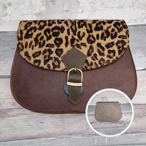 Puede incluir: Un bolso de cuero marrón con solapa de estampado de leopardo, una correa central de cuero marrón oscuro con hebilla de metal dorado y un recuadro circular que muestra la parte trasera lisa del bolso con un borde de estampado de leopardo.