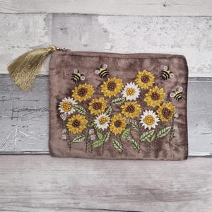 Könnte beinhalten: Eine braune Samt-Reißverschlusstasche mit einem gestickten Design aus Sonnenblumen und Bienen. Die Sonnenblumen sind gelb und weiß mit grünen Blättern. Eine goldene Quaste hängt am Reißverschluss.