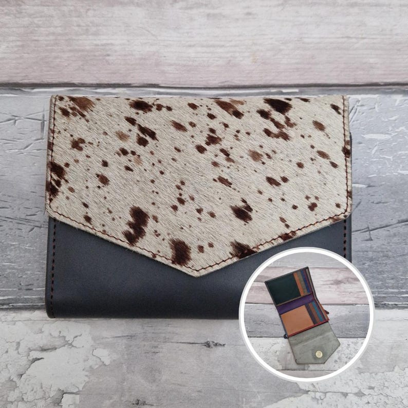 May include: A bi-fold wallet with a cowhide flap and a dark grey leather body. The flap has a light tan and brown spotted pattern. An inset shows the wallet open, revealing multiple compartments.