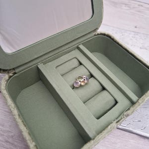 May include: Open sage green jewelry box with a built-in mirror. Inside, a silver ring with multiple colorful gemstones rests on a padded section. The ring features a cluster of light pink, green, and purple stones.