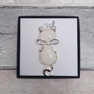 May include: A silver-toned cat-shaped brooch featuring a white pearlescent inlay in the head and body, accented by a small bow at the neck and a curled tail. The brooch is displayed on a white card with the text Sassy Spirit printed in black.