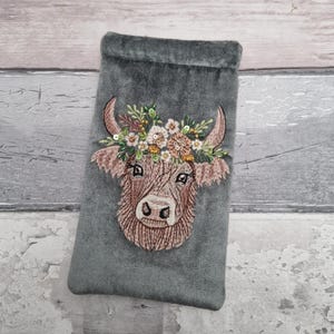 Puede incluir: Bolsa de terciopelo gris con un bordado de una vaca de las Tierras Altas con una corona de flores. La vaca es marrón y beige, con una corona de flores multicolor. La bolsa es rectangular y tiene una textura suave y aterciopelada.