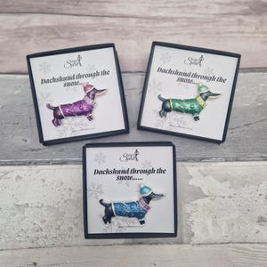 Può includere: Tre spille a forma di bassotto in scatole. Ogni spilla raffigura un bassotto con un maglione e un cappello, con la scritta "Dachshund through the snow..." sulla scatola. Le spille sono viola, verdi e blu.