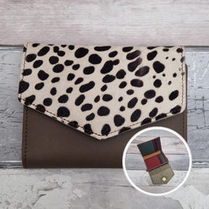 May include: A brown leather wallet with a flap closure. The flap features a cream-coloured material with black spots. The wallet has a small, circular inset showing the interior with multiple compartments.