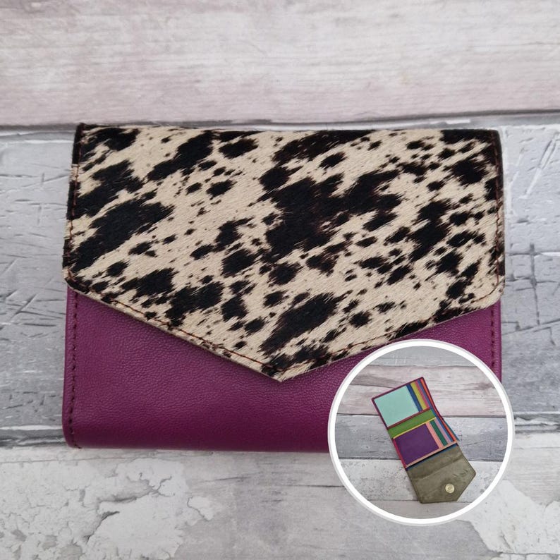 May include: A purple leather wallet with a cowhide flap featuring black and tan spots. The wallet has a snap closure and a small, colourful interior. The wallet is rectangular and appears to be new.