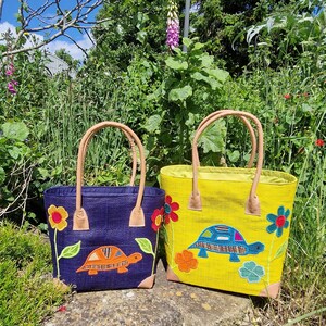 May include: Two tote bags, one navy blue and one yellow, with turtle and flower appliqués. The bags have tan handles and corner accents. The navy bag has an orange turtle, and the yellow bag has a blue turtle. The bags are sitting on a stone surface.