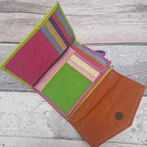 May include: A multi-coloured leather wallet, open to display its contents. The wallet features a bright palette of pink, green, orange, and yellow. It has several compartments, including card slots and a coin pouch. The wallet is open, showing its interior.