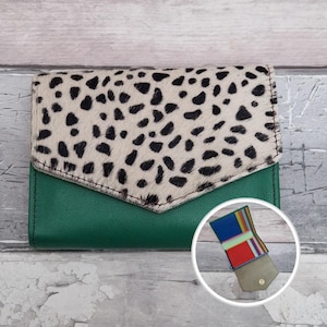 May include: A green leather wallet with a flap closure. The flap features a cream-coloured material with black spots. The wallet has a small, open compartment with colourful stripes. The wallet is on a wooden surface.