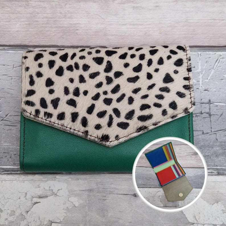 May include: A green leather wallet with a flap closure. The flap features a cream-coloured material with black spots. The wallet has a small, open compartment with colourful stripes. The wallet is on a wooden surface.