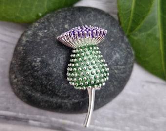 Puede incluir: Broche de cardo plateado con una flor morada y un cuerpo verde. El broche presenta pequeños detalles plateados y un largo tallo. El cardo está sobre una piedra gris oscura.