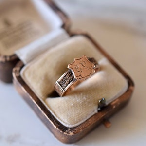 Op de afbeelding: Een gouden ring met een gegraveerd schildvormig ontwerp. Het schild heeft de initialen "M.R." gegraveerd. De ring zit in een klein, bruin fluwelen doosje.