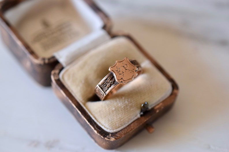 Op de afbeelding: Een gouden ring met een gegraveerd schildvormig ontwerp. Het schild heeft de initialen "M.R." gegraveerd. De ring zit in een klein, bruin fluwelen doosje.