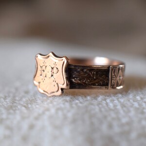 Op de afbeelding: Een gouden ring met een schildvormig ontwerp met de initialen "H.P." De ringband is versierd met een geweven patroon en een geometrisch ontwerp.