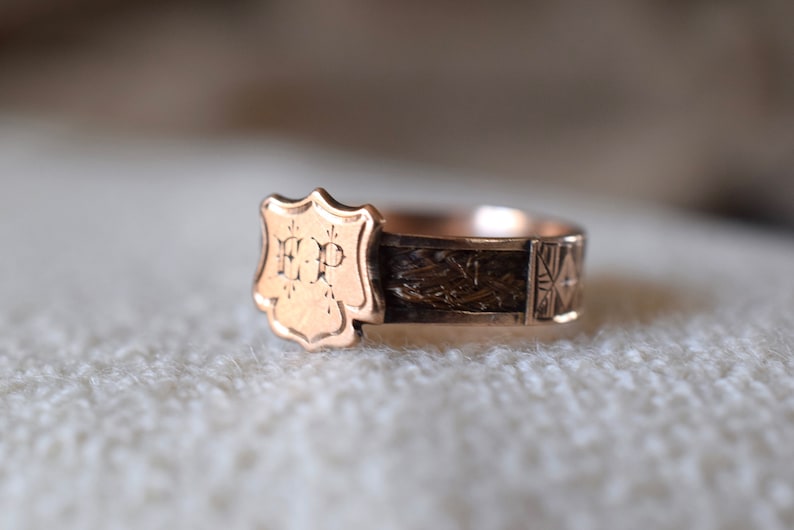 Op de afbeelding: Een gouden ring met een schildvormig ontwerp met de initialen "H.P." De ringband is versierd met een geweven patroon en een geometrisch ontwerp.