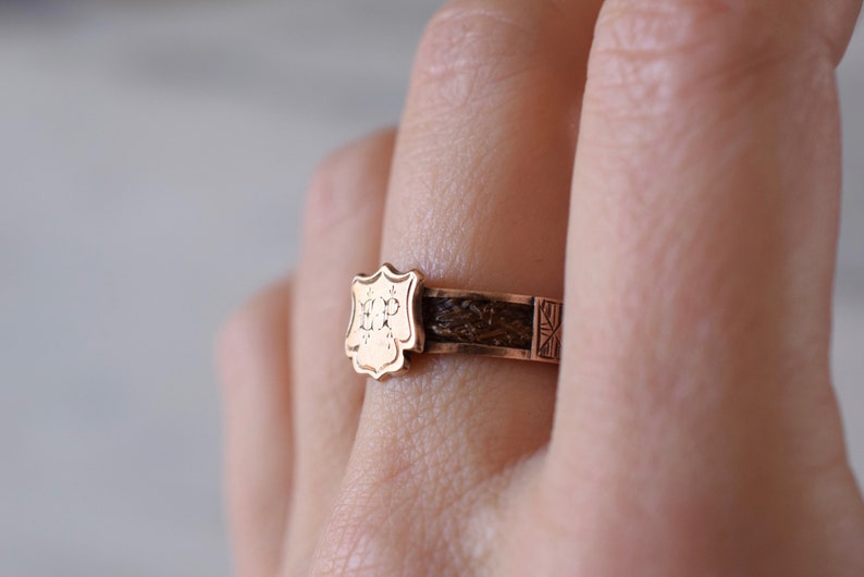 Op de afbeelding: Een gouden ring met een schildvormig ontwerp met de initialen "HP" gegraveerd aan de voorkant. De band van de ring is gemaakt van bruin gevlochten haar.