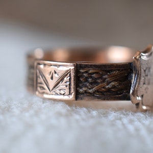 Op de afbeelding: Een gouden ring met een geweven haarontwerp en een schildvormige hanger gegraveerd met de initialen "M.M."