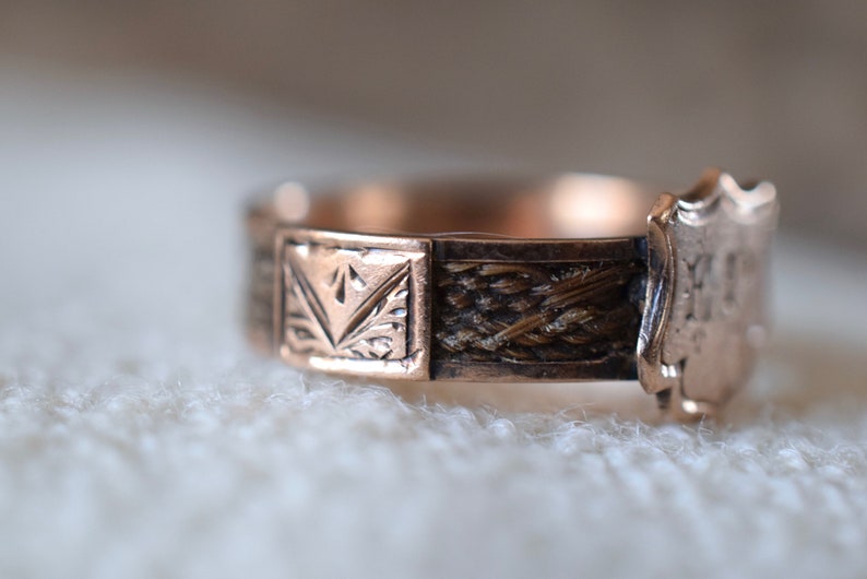 Op de afbeelding: Een gouden ring met een geweven haarontwerp en een schildvormige hanger gegraveerd met de initialen "M.M."