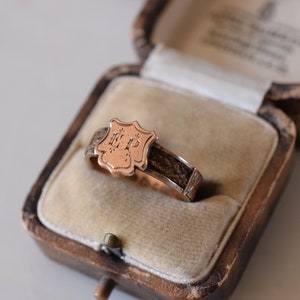 Op de afbeelding: Een gouden ring met een schildvormig ontwerp met de initialen "E.P." in een kleiner lettertype. De ring bevindt zich in een fluwelen doos.
