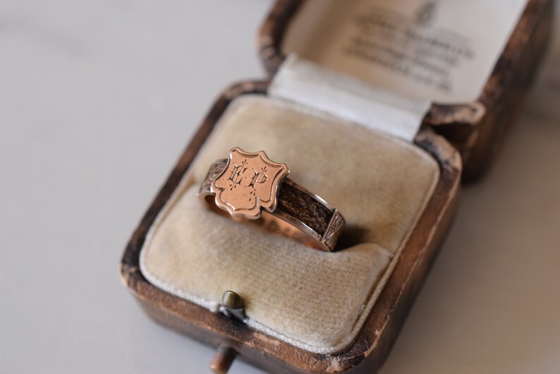 Op de afbeelding: Een gouden ring met een schildvormig ontwerp met de initialen "E.P." in een kleiner lettertype. De ring bevindt zich in een fluwelen doos.