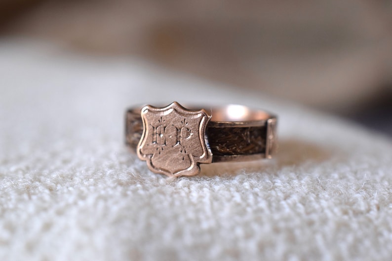 Op de afbeelding: Een gouden ring met een schildvormig ontwerp met de initialen "H.P." gegraveerd aan de voorkant. De band van de ring is gemaakt van geweven haar.