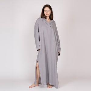 May include: A light grey hooded maxi dress with long sleeves and a side split. The dress has a relaxed fit and a V-neckline. The model is wearing the dress in a studio setting.