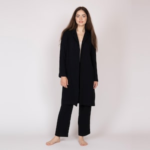 Puede incluir: Un abrigo largo negro y pantalones negros a juego. El abrigo tiene cuello con solapas y una silueta larga y recta. Los pantalones son de pierna ancha. El atuendo lo lleva una persona de pie frente a un fondo blanco.