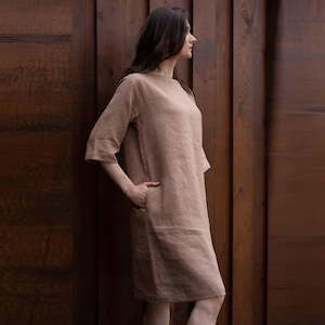 May include: A knee-length dress in a solid light brown color, featuring a round neckline, three-quarter length sleeves, and side pockets. The garment has a straight, relaxed fit and is constructed from a textured fabric.