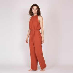 Könnte beinhalten: Ein rostfarbener Jumpsuit mit Neckholder-Ausschnitt und weitem Bein. Das einteilige Kleidungsstück hat eine geraffte Taille und Taschen. Das Model steht vor einem weißen Hintergrund. Der Jumpsuit ist aus leichtem Stoff.