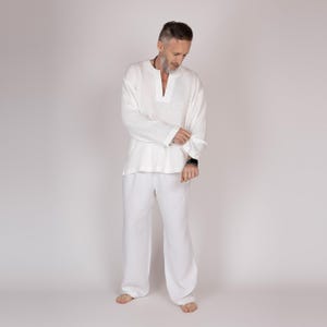 May include: A white linen outfit, comprising a long-sleeved tunic with a V-neck and matching wide-leg trousers. The outfit is made of a lightweight fabric. The model is wearing the outfit.
