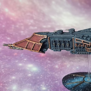 Può includere: Un modello dettagliato di navicella spaziale in miniatura, prevalentemente blu scuro con accenti color rame. Il modello presenta dettagli intricati, tra cui torrette e pannelli, ed è montato su un supporto trasparente.