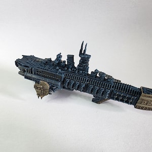 Puede incluir: Modelo detallado de un buque de guerra en miniatura en tonos azules y beige. El modelo presenta detalles intrincados, incluyendo torretas, múltiples niveles y un casco texturizado. El barco está diseñado para juegos de mesa o exhibición.