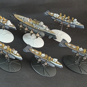 May include: Six miniature model warships. The ships are painted in shades of grey, brown, and gold, with detailed features like turrets and smokestacks. Each ship is mounted on a stand, some with circular bases and others with oval bases.