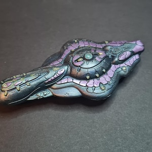 Puede incluir: Una escultura metálica detallada con un diseño futurista. El objeto presenta una base gris oscuro con acentos morados y turquesas. El intrincado diseño incluye patrones circulares y lineales, lo que sugiere un tema tecnológico o de ciencia ficción.