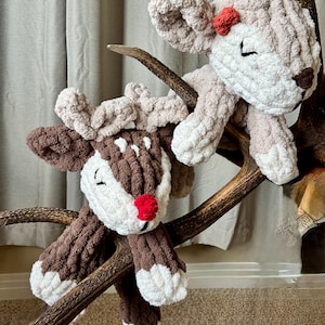 May include: Two plush reindeer toys, one brown and white and one beige, are perched on a large antler. The brown reindeer has a red nose and the beige reindeer has a red bow.