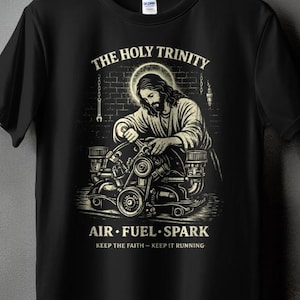 May include: Black t-shirt with a graphic of Jesus working on an engine, with the text "THE HOLY TRINITY" above and "AIR • FUEL • SPARK" below. The phrase "KEEP THE FAITH - KEEP IT RUNNING" is also included.