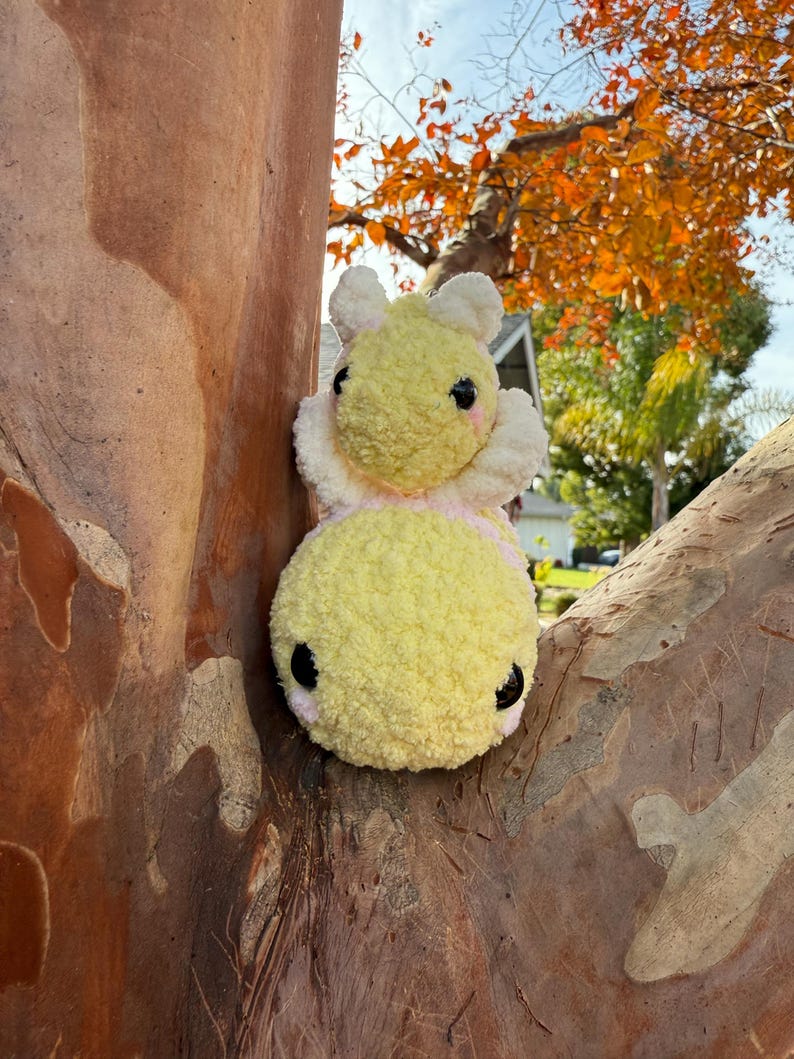 May include: A handmade, plush bee toy with a yellow body and white wings. The bee has black eyes and pink cheeks. The toy is made of soft, textured yarn and is designed to be a cute and cuddly item.